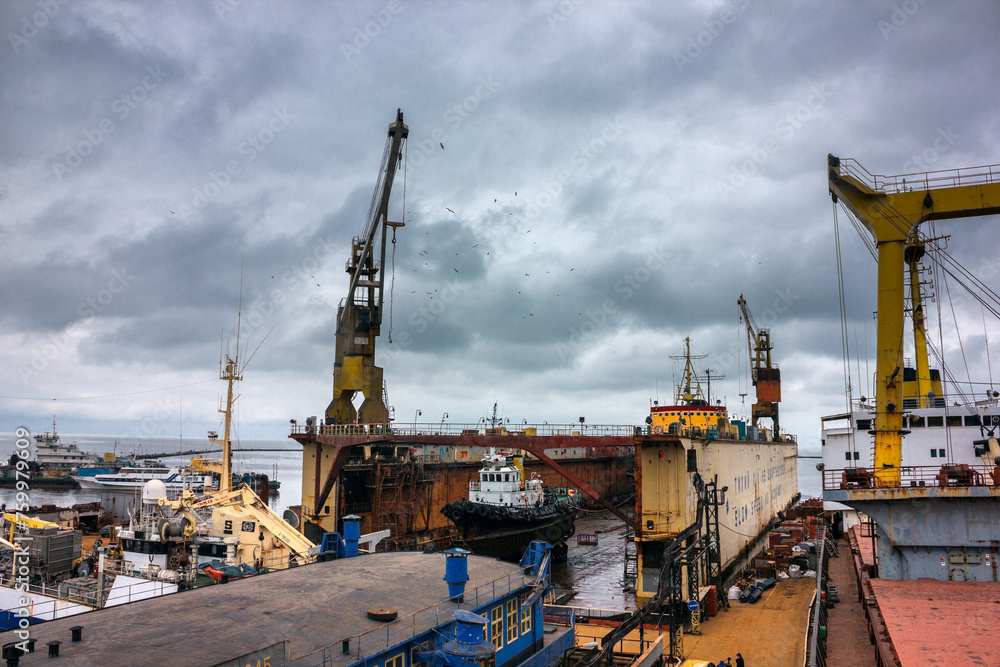 Industrial ship repair yard with floating dock, vessels undergo maintenance. Tugboats assist in marine operations, heavy cranes lift equipment. Overcast sky over maritime workplace, shipping industry