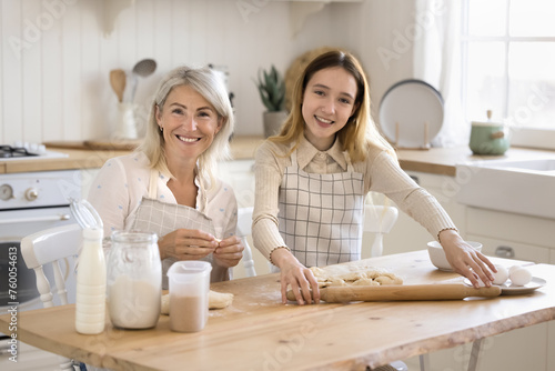 Beautiful middle-aged woman preparing pastries with cute teenager daughter in the kitchen, flattening homemade dough for holiday buns, teaching preteen girl to cook at home. Culinary, family recipe