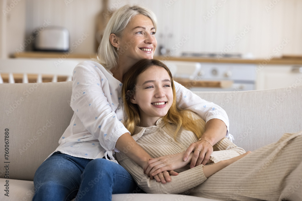 Cheerful adolescent girl spends leisure time with mature mother at home, sit resting on sofa together, enjoy tender moments, show love and care. Happy family of two generations hugging and daydreaming