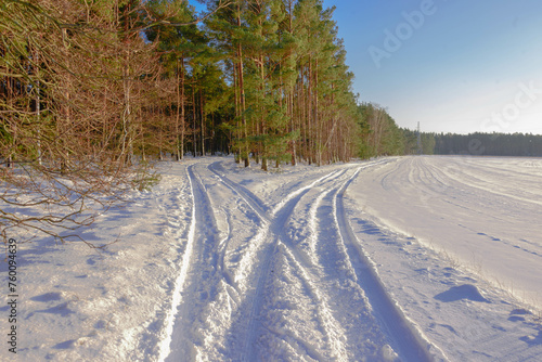 Równina pokryta warstwą śniegu. Jest słoneczny, bezchmurny dzień. W śniegu odciśnięte są ślady kół samochodowych tworzące rozwidlenie, z których lewe skręca do lasu, prawe w okoliczne pola.