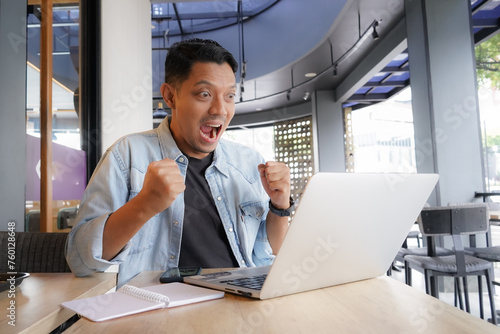 Asian man happy and enthusiastic with blue shirt using laptop and mobile phone in coffee shop, online shopping business