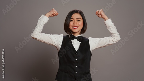 Asian hotel concierge flexing arms muscles in grey studio, showing off her strength and power in front of camera. Receptionist with bow and uniform standing with biceps flexed. Camera B.
