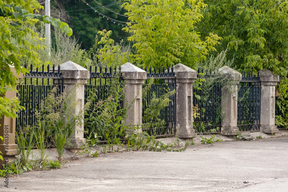 Old stone columns and an iron lattice of an abandoned ancient fence, stands along an asphalt road, overgrown with green bushes and grass, an abandoned yard Old stone columns and an iron lattice of an abandoned ancient fence, stands along an asphalt road, overgrown with green bushes and grass, an abandoned yard