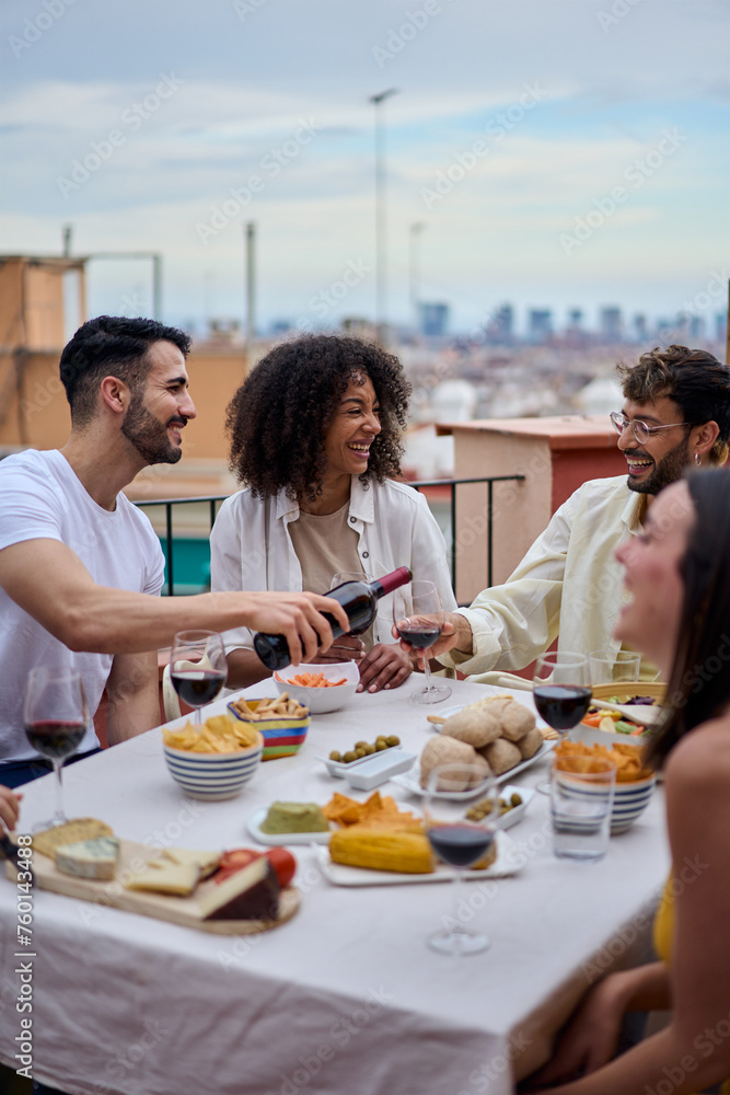 Vertical young smiling Caucasian man serving red wine to guests at food table in celebration with happy friends on rooftop. Group of cheerful friends gathered for lunch party on outdoors. Copy space