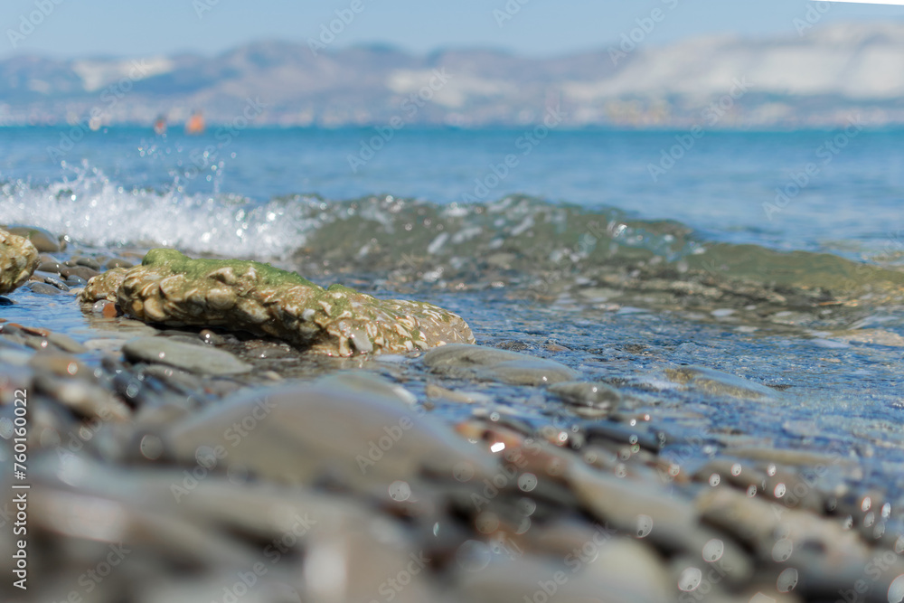 Seascape - wet round pebbles in the foreground are covered with a wave of the blue sea, mountain hills are visible in the distance on the horizon
