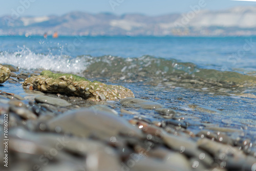 Obraz na płótnie Seascape - wet round pebbles in the foreground are covered with a wave of the blue sea, mountain hills are visible in the distance on the horizon w salonie