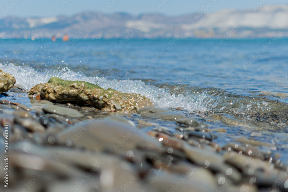 Seascape - wet round pebbles in the foreground are covered with a wave of the blue sea, mountain hills are visible in the distance on the horizon