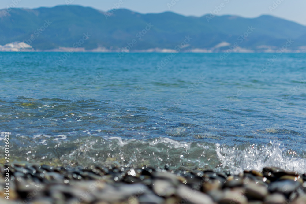Seascape - wet round pebbles in the foreground are covered with a wave of the blue sea, mountain hills are visible in the distance on the horizon