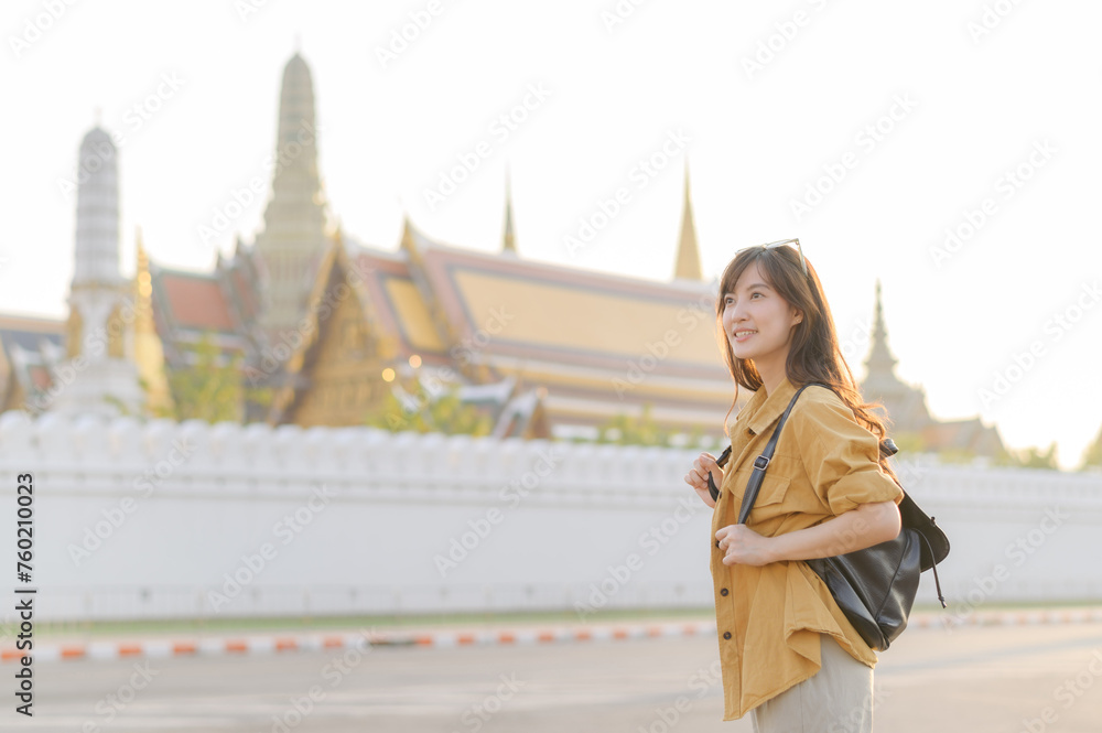 Traveler asian woman in her 30s, backpack slung over her shoulder, explores the intricate details of Wat Pra Kaew with childlike wonder. Sunlight dances on the golden rooftops. Traveler asian woman in her 30s, backpack slung over her shoulder, explores the intricate details of Wat Pra Kaew with childlike wonder. Sunlight dances on the golden rooftops.
