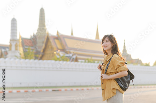 Traveler asian woman in her 30s, backpack slung over her shoulder, explores the intricate details of Wat Pra Kaew with childlike wonder. Sunlight dances on the golden rooftops. © Jirawatfoto