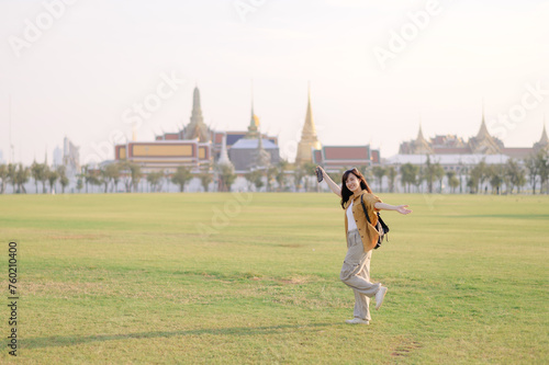Traveler asian woman in her 30s, backpack slung over her shoulder, explores Wat Pra Kaew with childlike wonder. Sunlight dances on the golden rooftops, and a carefree smile