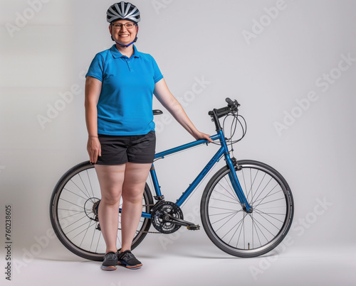 A woman in a blue polo shirt and black shorts stands next to her blue bicycle in a studio photo, wearing a bicycle helmet.