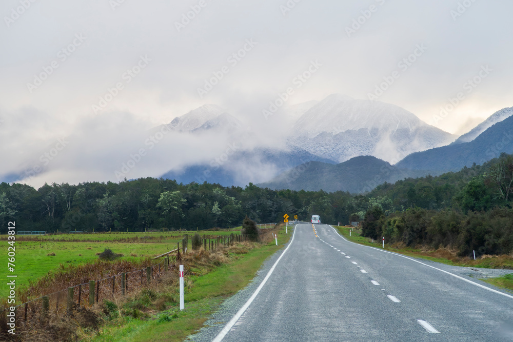 Photograph of a van with tourists driving on a road towards a mountain range covered in snow and mist in Fiordland National Park on the South Island of New Zealand Photograph of a van with tourists driving on a road towards a mountain range covered in snow and mist in Fiordland National Park on the South Island of New Zealand