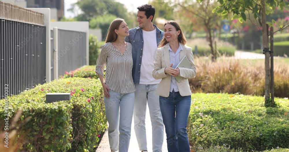 Real-estate agent and young couple walking along well-groomed garden in closed residential complex with its own territory, lush green area and entering house. Property buyers at meeting with realtor Real-estate agent and young couple walking along well-groomed garden in closed residential complex with its own territory, lush green area and entering house. Property buyers at meeting with realtor