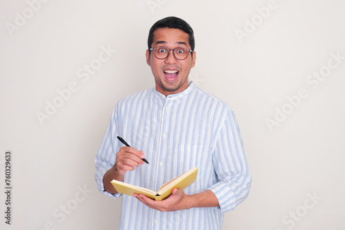 Adult Asian man showing wow face expression when holding a book and pen photo