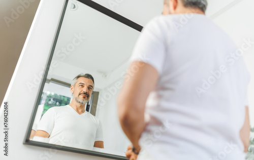 Confident mature man in white T-shirt admiring his reflection, showcasing self-assurance and personal care