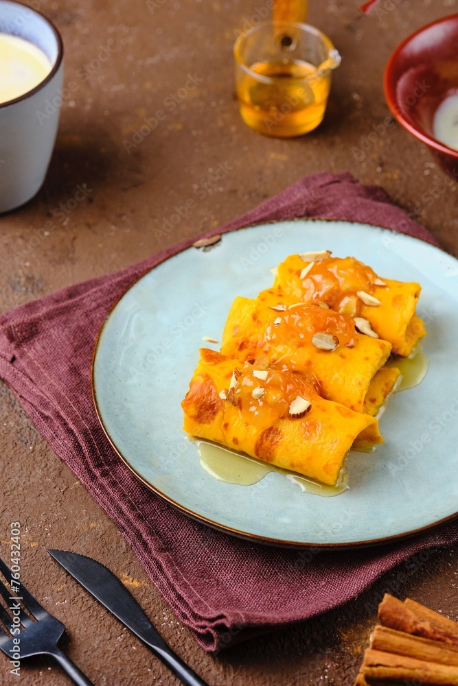 Thin pumpkin pancakes rolled into a roll and topped with honey and jam on a gray ceramic plate on a brown concrete background. Pancake recipes. Thin pumpkin pancakes rolled into a roll and topped with honey and jam on a gray ceramic plate on a brown concrete background. Pancake recipes.