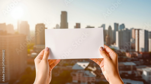Woman's hands holding a blank white card against the backdrop of a metropolis with high-rise buildings. Layout concept for a postcard to a business meeting, event or vacation. Generated AI.