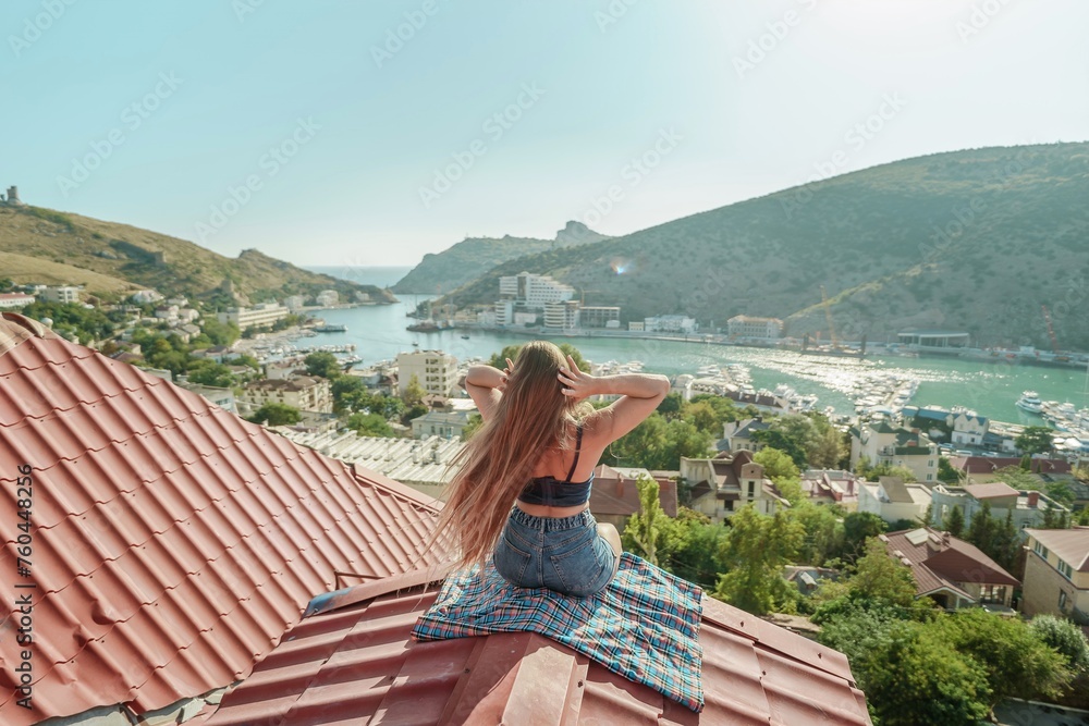 Woman sits on rooftop, enjoys town view and sea mountains. Peaceful rooftop relaxation. Below her, there is a town with several boats visible in the water. Rooftop vantage point.