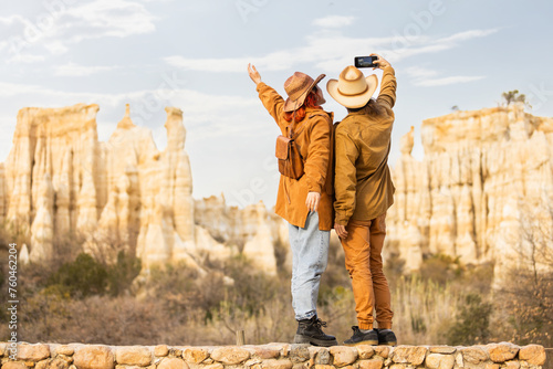 A couple is taking a picture of a mountain range. The man is holding a cell phone and the woman is wearing a hat