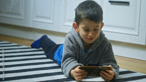 Cute little boy laying on the floor in kitchen playing mobile game on smartphone at home. Kid using phone for gaming. 