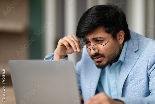 Indian businessman squinting through eyeglasses looking at laptop in office