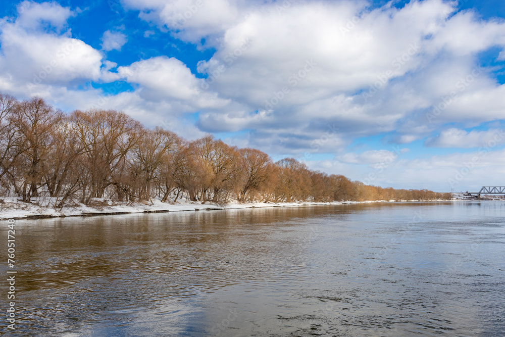 Trees along the river, clouds reflected in the water, spring on the river, salty weather, high water, view from the middle of the river to the shore. Blue sky with clouds