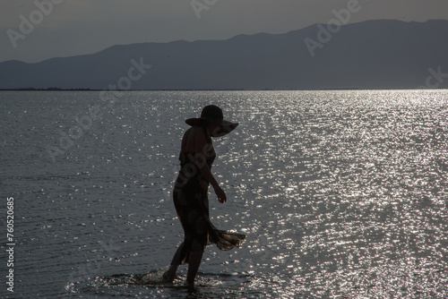Fototapeta Naklejka Na Ścianę i Meble -  Silueta de mujer al anochecer, caminando por dentro del agua en la playa, vestida y con pamela 