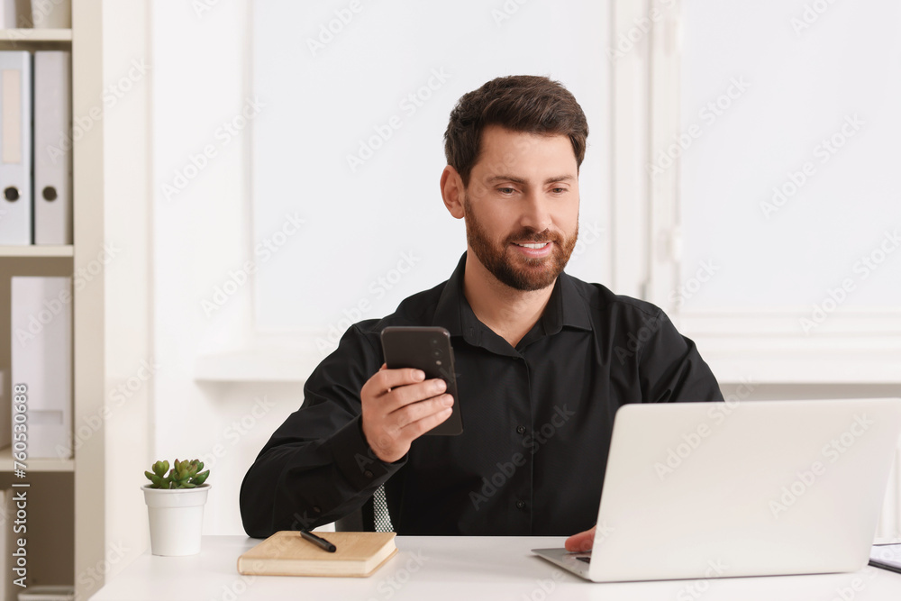 Smiling man with smartphone using laptop at table in office Smiling man with smartphone using laptop at table in office