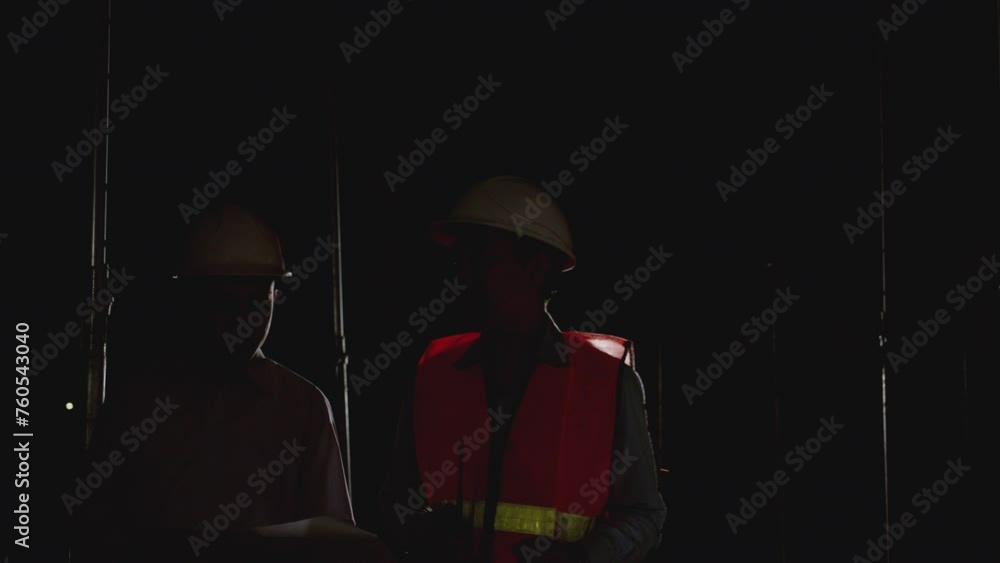 Working Late at Night in construction site. Engineers and architects in safety uniforms and hard hats talk and review blueprints as they walking in a building site. Working Late at Night in construction site. Engineers and architects in safety uniforms and hard hats talk and review blueprints as they walking in a building site.