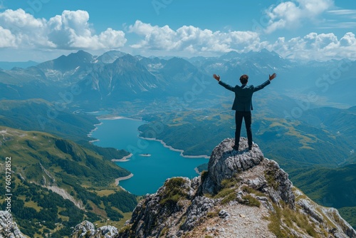 A businessman stands on a rocky mountain peak with arms raised in victory, overlooking a stunning landscape with a serpentine lake nestled among lush green valleys and rugged mountain ranges