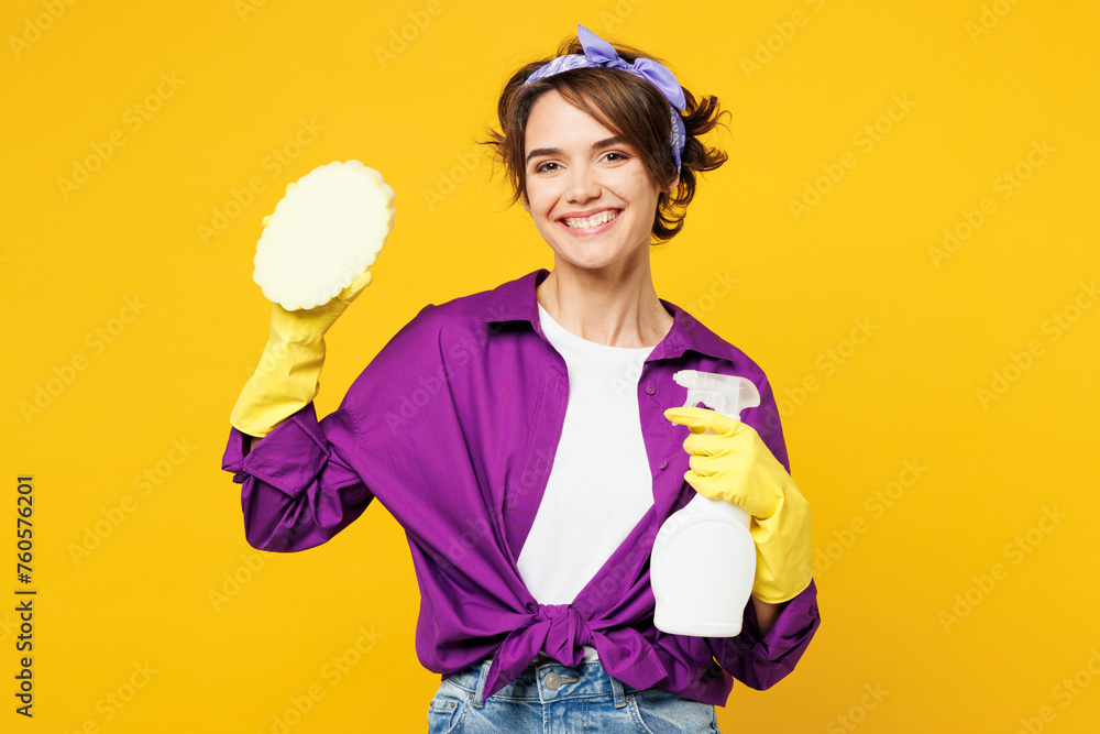 Young happy smiling woman wears purple shirt rubber gloves do housework tidy up hold in hand sponge spray bottle look camera isolated on plain yellow background studio portrait. Housekeeping concept. Young happy smiling woman wears purple shirt rubber gloves do housework tidy up hold in hand sponge spray bottle look camera isolated on plain yellow background studio portrait. Housekeeping concept.
