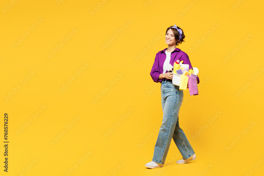 Fototapeta premium Full body sideways young smiling happy woman wear casual purple shirt do housework tidy up hold basin with laundry clothes go isolated on plain yellow background studio portrait. Housekeeping concept.