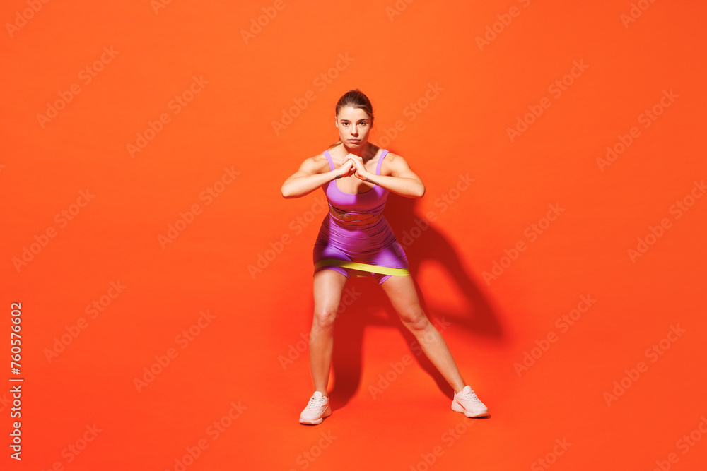 Full body young fitness trainer instructor woman sportsman wearing purple top clothes in home gym hold elastic rubber band for legs isolated on plain orange background. Workout sport fit abs concept.