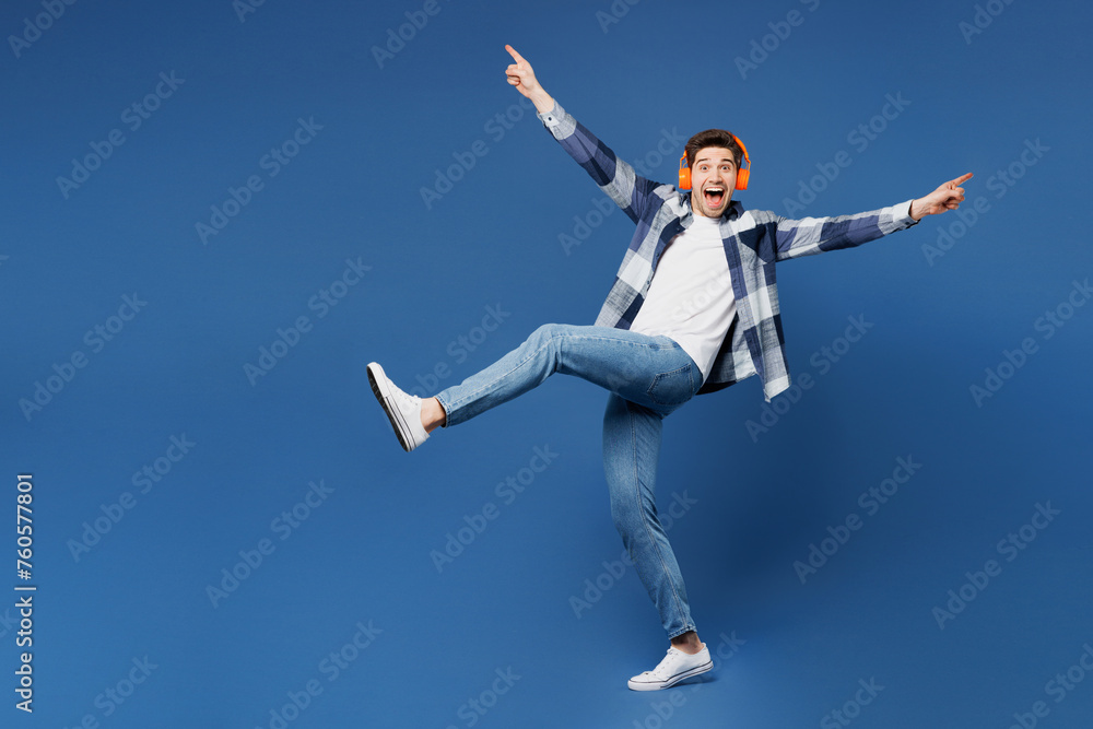 Full body young happy man wear shirt white t-shirt casual clothes listen to music in headphones with outstretched hands legs isolated on plain blue cyan background studio portrait. Lifestyle concept.