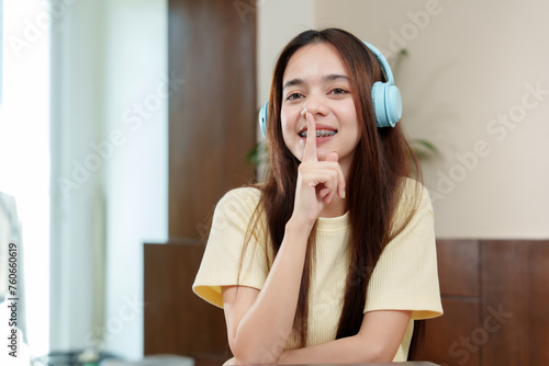 Asian lady with long hair, braces, wearing headset, gestures for silence, finger on lips, eyes gleaming with playful secrecy. Mirthful expression on face of person with dental braces, signalling hush