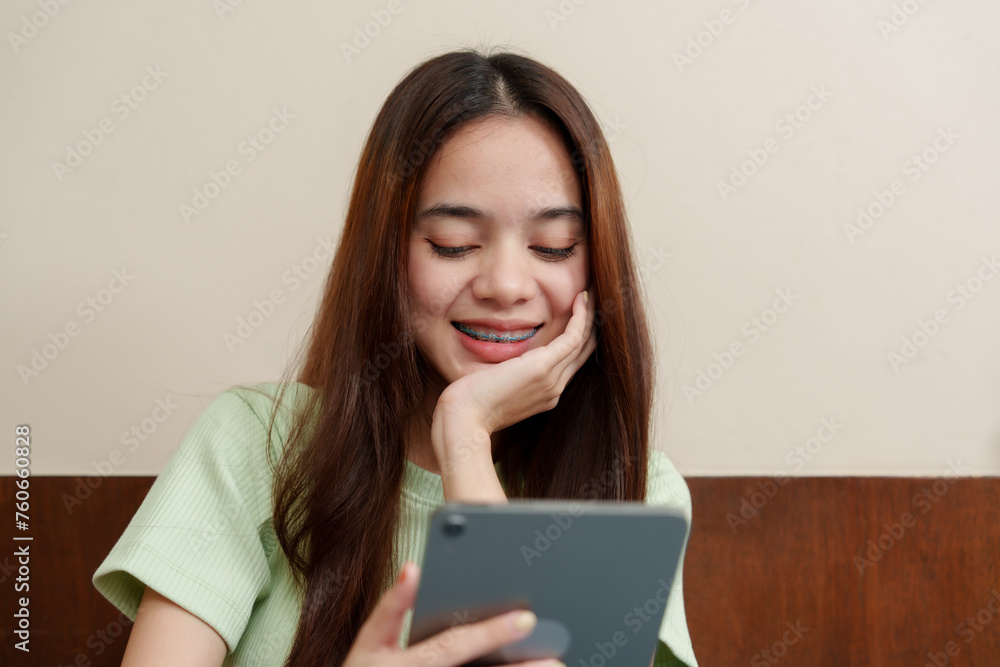 Radiant Asian student. woman gazes at a tablet screen, content in her at-home learning setup, brimming with eagerness. Serene learner looks at her device screen, settled in her personal study area Radiant Asian student. woman gazes at a tablet screen, content in her at-home learning setup, brimming with eagerness. Serene learner looks at her device screen, settled in her personal study area