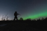 A military soldier with a night vision device and a rifle with a suppressor in the forest against the background of the starry sky and northern lights.