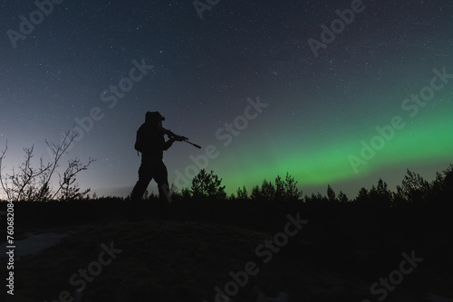 A military soldier with a night vision device and a rifle with a suppressor in the forest against the background of the starry sky and northern lights. photo