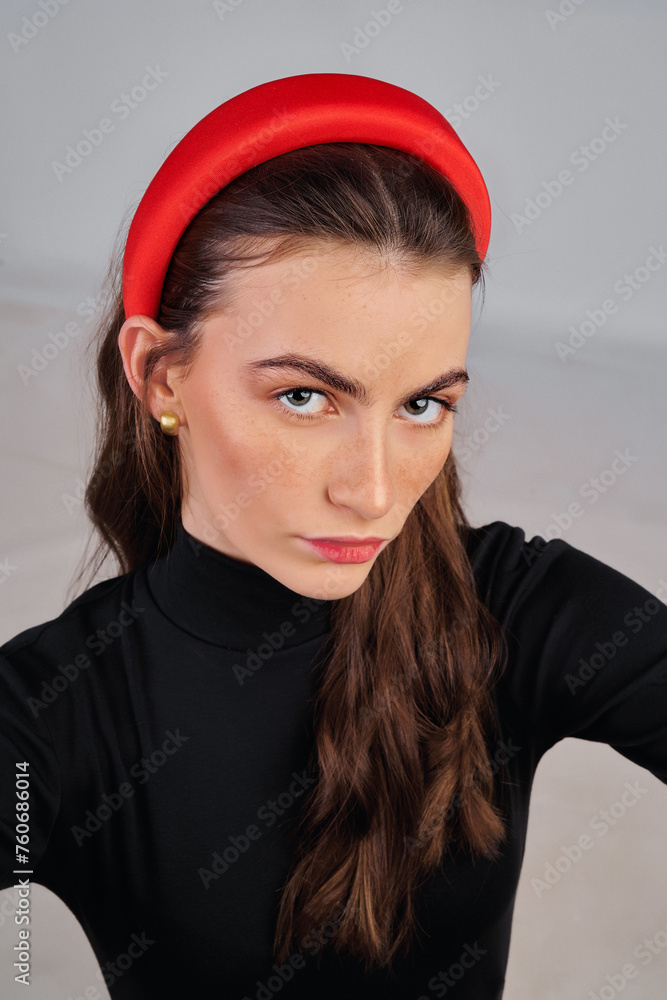 Close-up shot of a young beautiful woman girl with loose hair wearing a black turtleneck and a red headband. European girl in a fashionable wide headband taking selfies against a gray background. Close-up shot of a young beautiful woman girl with loose hair wearing a black turtleneck and a red headband. European girl in a fashionable wide headband taking selfies against a gray background.