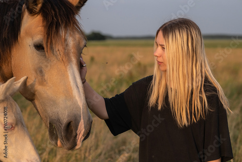 A young beautiful blonde girl plays with a horse on the field.
