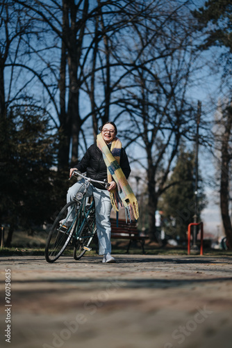 Joyful middle-aged woman wearing glasses and a colorful scarf, standing with her bike in a sunny park setting, exuding positivity and an active lifestyle. © qunica.com