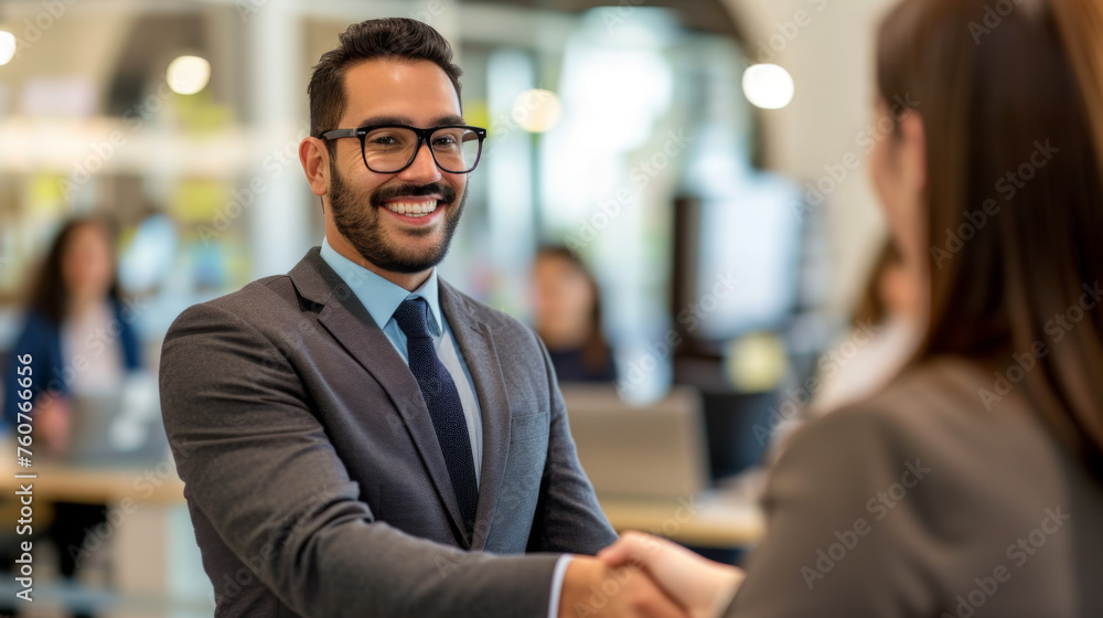 Fototapeta premium A bearded man in a suit with glasses is smiling and shaking hands with a colleague in a modern office environment.