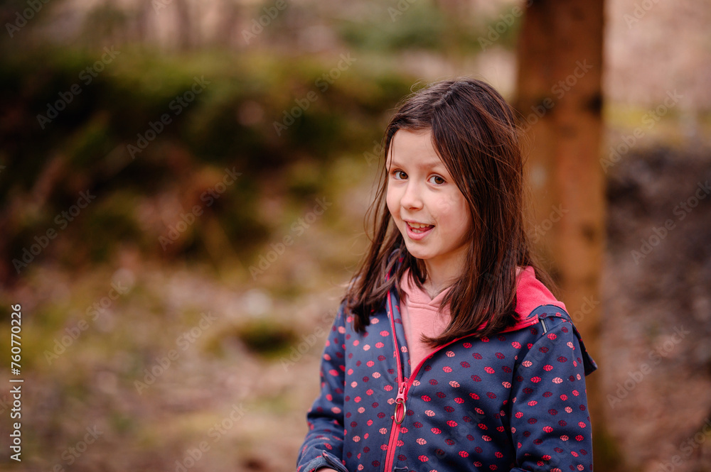 A cheerful young girl with brown hair smiles brightly, wearing a dotted jacket in a lush forest setting, exuding warmth and innocence A cheerful young girl with brown hair smiles brightly, wearing a dotted jacket in a lush forest setting, exuding warmth and innocence