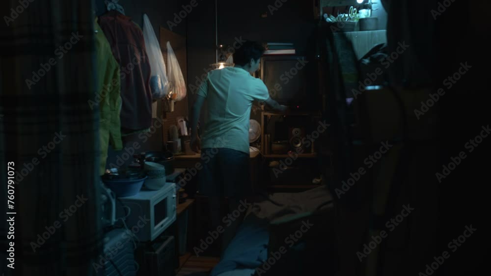 Full shot of young Asian man walking into micro apartment, switching on light bulb, turning on TV, laying down on bunk bed, pulling over blanket and watching movie, while resting at home after work Full shot of young Asian man walking into micro apartment, switching on light bulb, turning on TV, laying down on bunk bed, pulling over blanket and watching movie, while resting at home after work