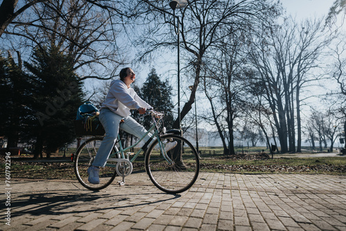 A joyful female cyclist with headphones on rides along a paved park path surrounded by bare trees and a clear sky.