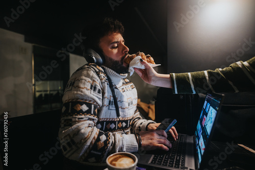 Focused man multitasking, having a snack while working on a laptop with headphones in a dimly lit room, capturing the essence of deadline pressure. photo