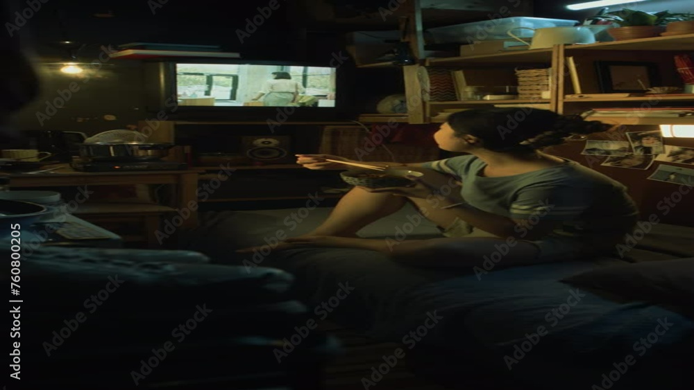 Full vertical shot of young Asian woman sitting with crossed legs on bed in cluttered tiny flat, eating stir-fry from bowl with chopsticks for dinner, watching beauty commercial or reality show on TV Full vertical shot of young Asian woman sitting with crossed legs on bed in cluttered tiny flat, eating stir-fry from bowl with chopsticks for dinner, watching beauty commercial or reality show on TV