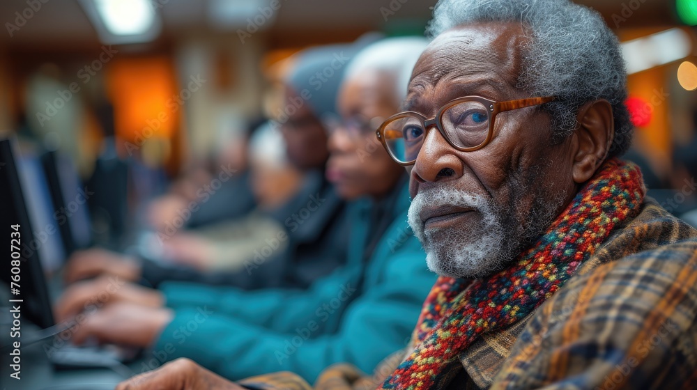 An elderly African American man with glasses and a scarf focuses intently while using a computer in a class.