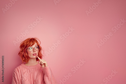 jeune femme rousse à taches de rousseurs, lunettes, qui doute et pose son index devant sa bouche pour montrer son hésitation, elle porte un pull en laine rose à torsades, sur fond rose avec copyspace photo